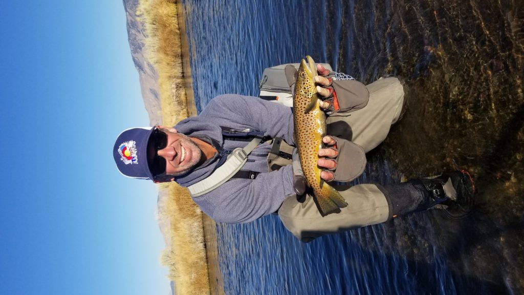 Fisherman with a hat holds up a large brown trout out of the over Owens River with grass on the sides and a large field