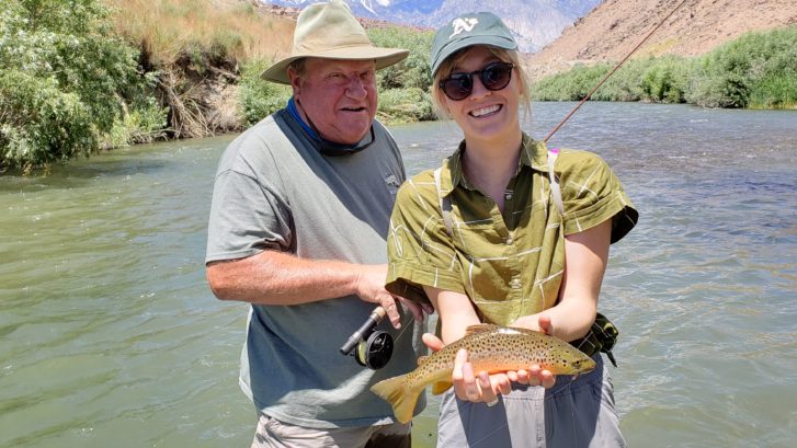 Father and daughter standing in the lower owens river while daughter holds up a 14" brown trout. Mountains in the background